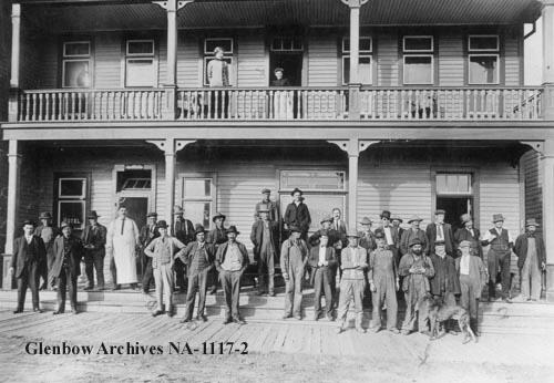 Exterior view of a building from across a dirt street. About 30 men are stood in front of the building posing for the photograph, which shows the verandah around the building and then a few steps down to a planked sidewalk laid on top of the dirt of the street.